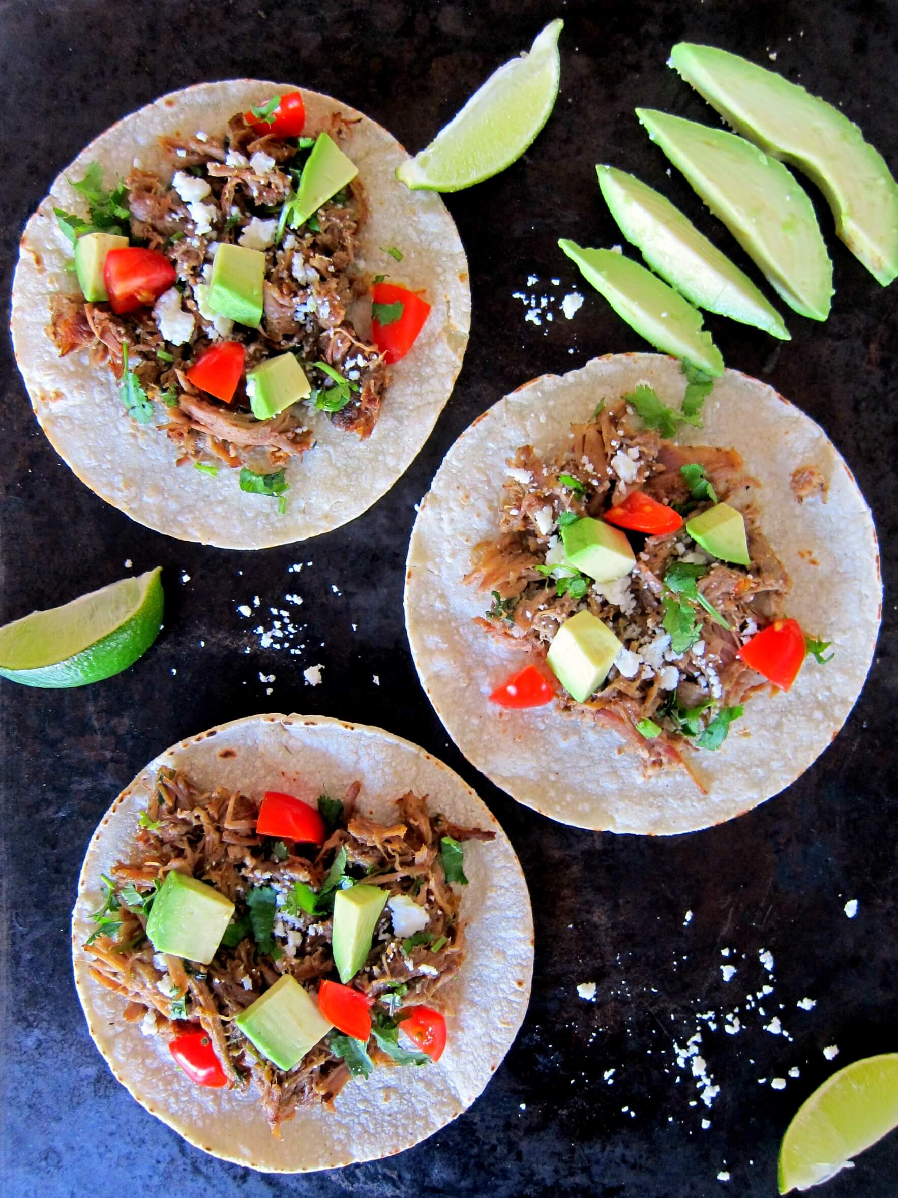 Instant Pot Carnitas open tacos topped with avocados and tomatoes on a black background - Paint the Kitchen Red