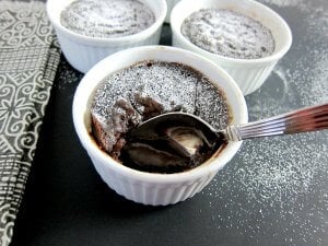 Instant Pot chocolate lava cake spoon into gooey center, with two intact lava cakes in background on black surface with powdered sugar dusting - Paint the Kitchen Red