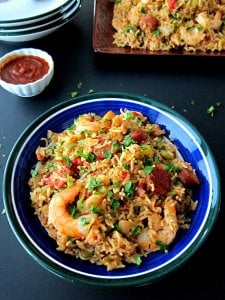 Instant Pot Jambalaya in blue bowl with hot sauce, stack of plates in background on a black table - Paint the Kitchen Red