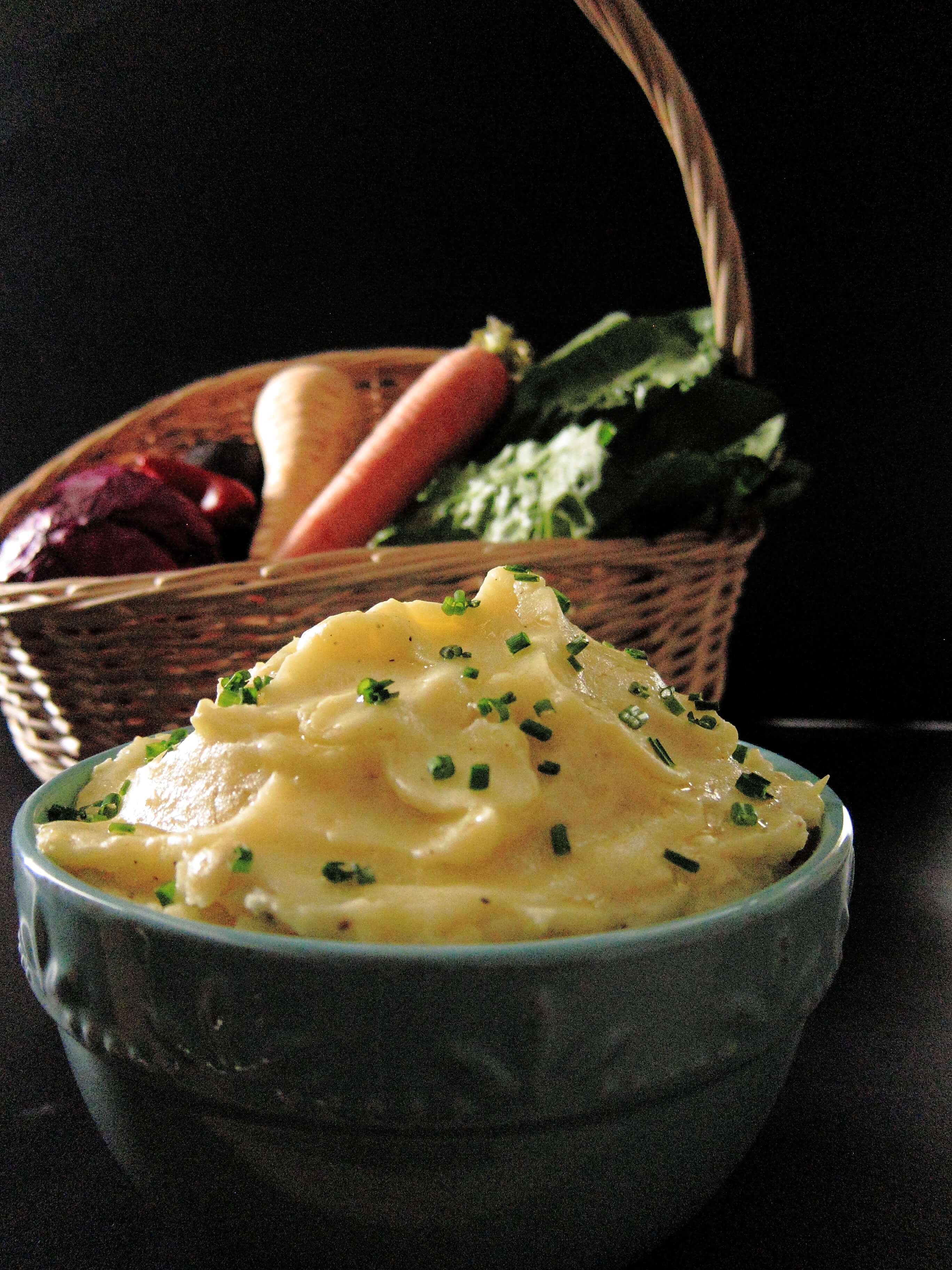 Bowl full of mashed parsnips and potatoes garnished with chives. A basket of vegetables in the background.