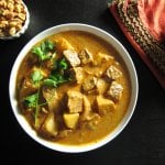 Instant Pot Beef Massaman Curry, golden colored and rich, in white bowl with bowl of peanuts and white rice on red and gold napkin on black background.