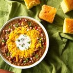 Instant Pot Chili in white bowl on green cloth with cornbread; topped with cheese, sour cream and green onions - Paint the Kitchen Red