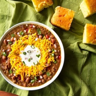 Instant Pot Chili in white bowl on green cloth with cornbread; topped with cheese, sour cream and green onions - Paint the Kitchen Red