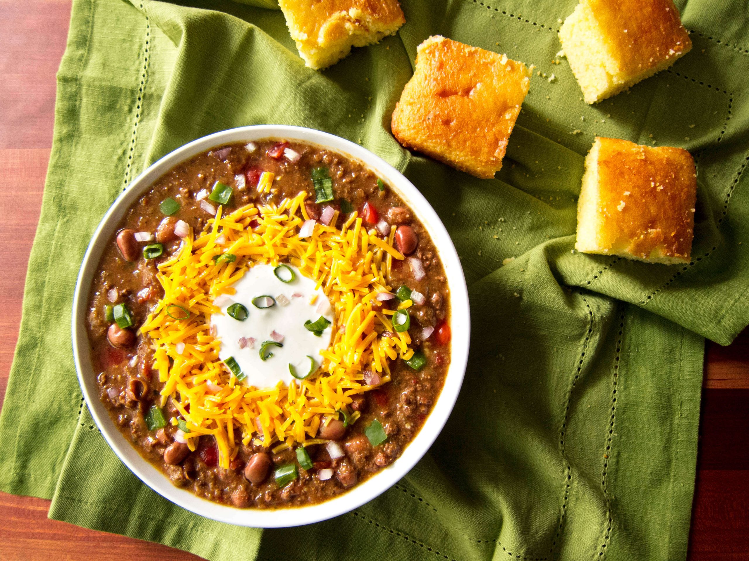 Instant Pot Chili in white bowl on green cloth with cornbread; topped with cheese, sour cream and green onions - Paint the Kitchen Red