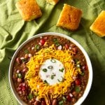 Instant Pot Chili in white bowl on green cloth with cornbread; topped with cheese, sour cream and green onions - Paint the Kitchen Red