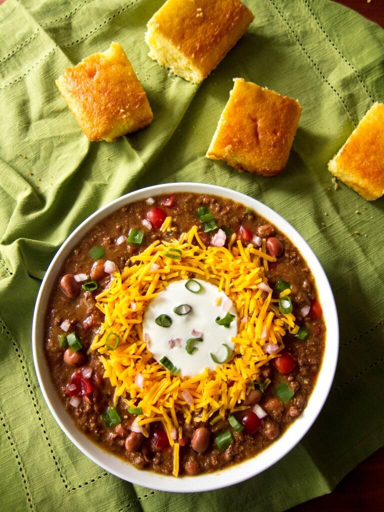 Instant Pot Chili in white bowl on green cloth with cornbread; topped with cheese, sour cream and green onions - Paint the Kitchen Red