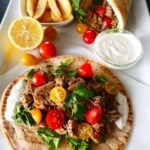 Instant Pot Chicken Shawarma - pita with white sauce, chicken, salad greens, halved cherry yellow and red tomatoes on parchment placed on a large white plate with French fries and white sauce and folded shawarma
