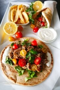 Instant Pot Chicken Shawarma - pita with white sauce, chicken, salad greens, halved cherry yellow and red tomatoes on parchment placed on a large white plate with French fries and white sauce and folded shawarma