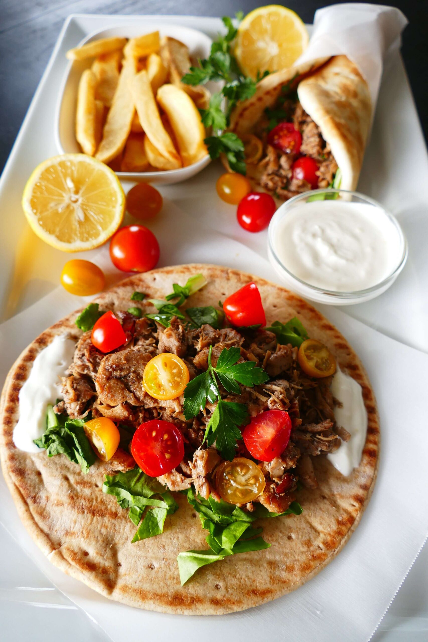 Instant Pot Chicken Shawarma - pita with white sauce, chicken, salad greens, halved cherry yellow and red tomatoes on parchment placed on a large white plate with French fries and white sauce and folded shawarma