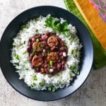 Instant Pot Red Beans and Sausage served on rice in a black bowl on a white wooden background with multicolor napkins. Garnished with parsley and green onions - Paint the Kitchen Red