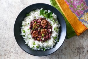 Instant Pot Red Beans and Sausage served on rice in a black bowl on a white wooden background with multicolor napkins. Garnished with parsley and green onions - Paint the Kitchen Red