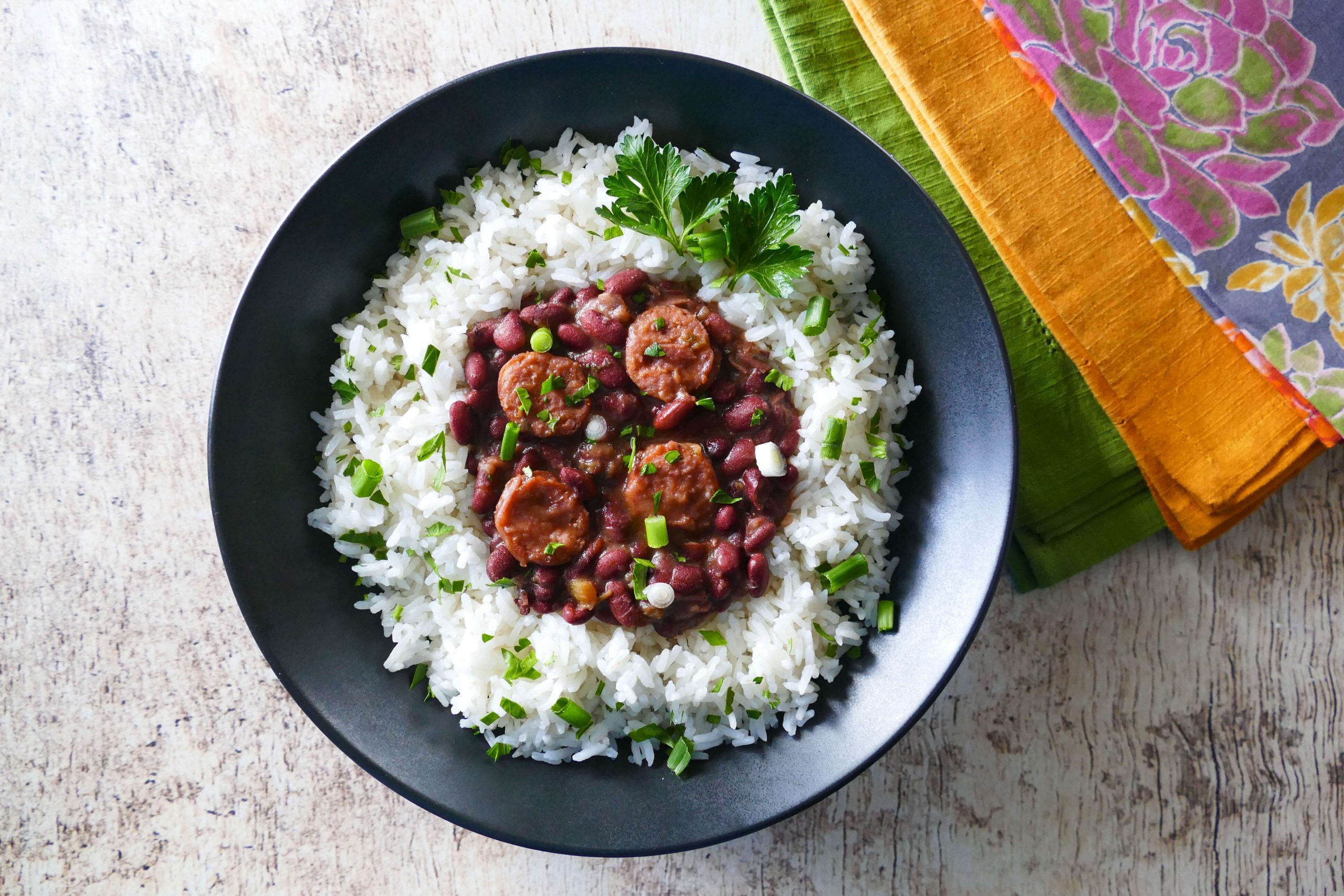 Instant Pot Red Beans and Sausage served on rice in a black bowl on a white wooden background with multicolor napkins. Garnished with parsley and green onions - Paint the Kitchen Red