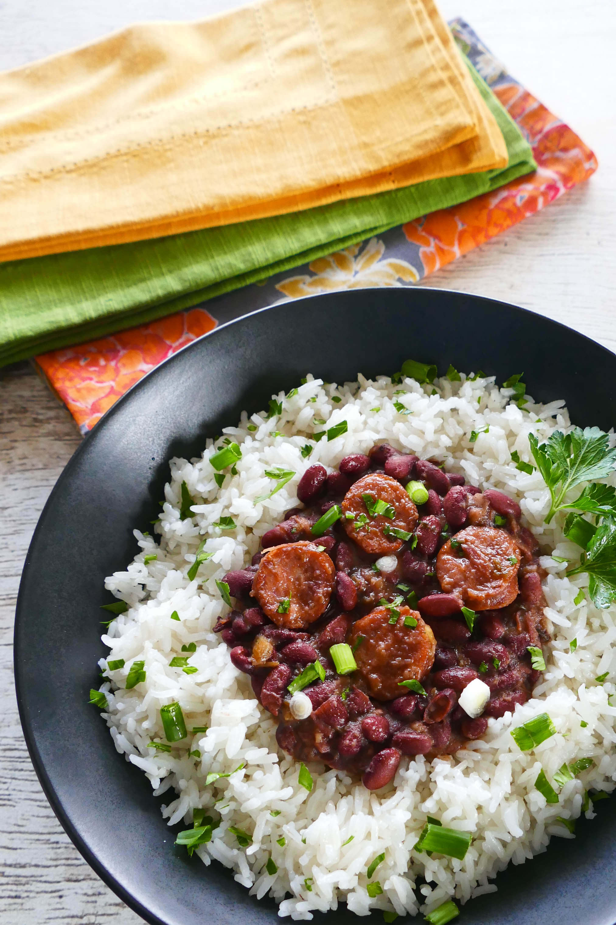 Instant Pot Red Beans and Rice - Beans and sausage served on rice in a black bowl on a white wooden background with multicolor napkins. Garnished with parsley and green onions - Paint the Kitchen Red