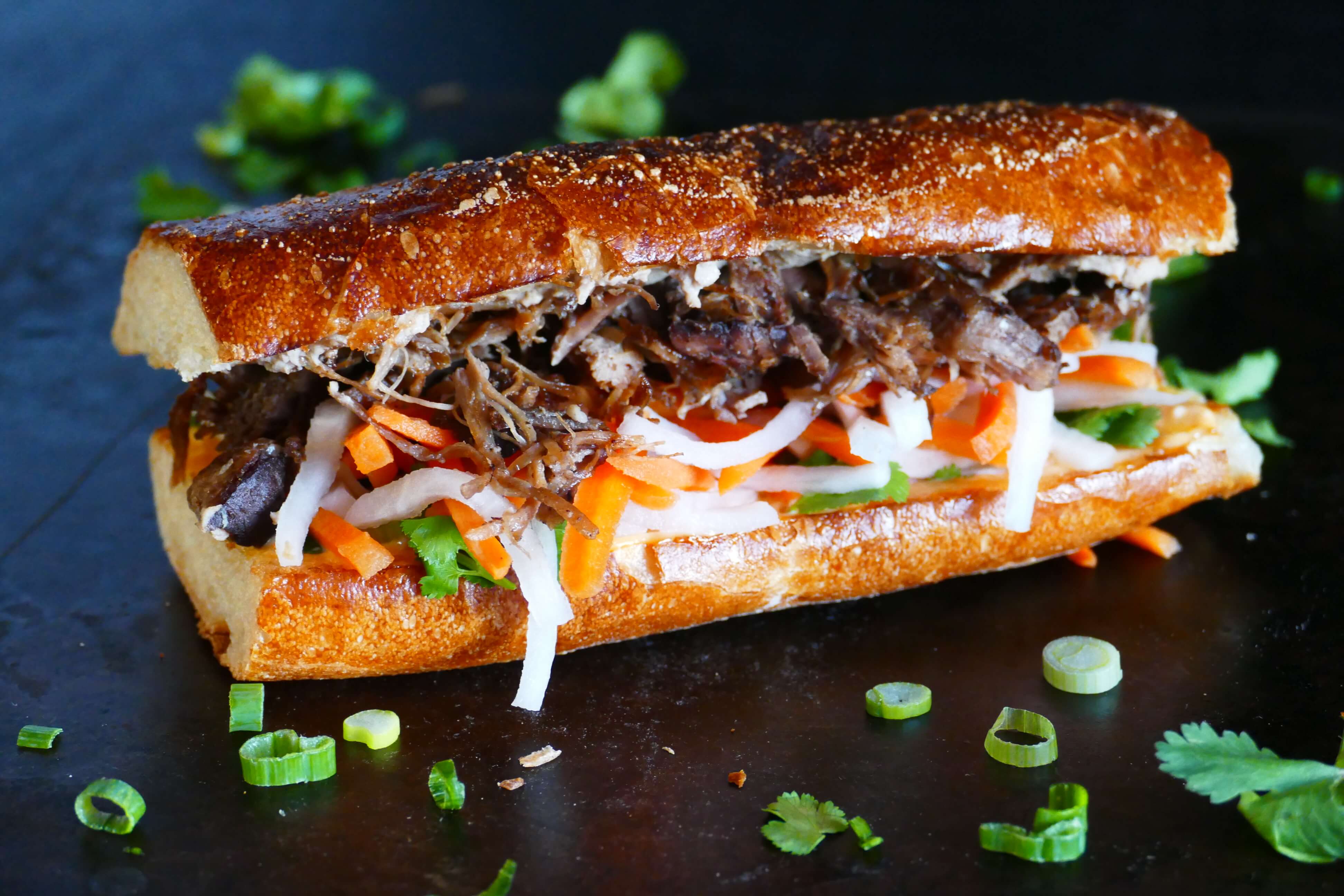 Instant Pot Banh Mi sandwich on black background, with meat, daikon, carrots and cilantro; cilantro and green onions sprinkled on surface - Paint the Kitchen Red