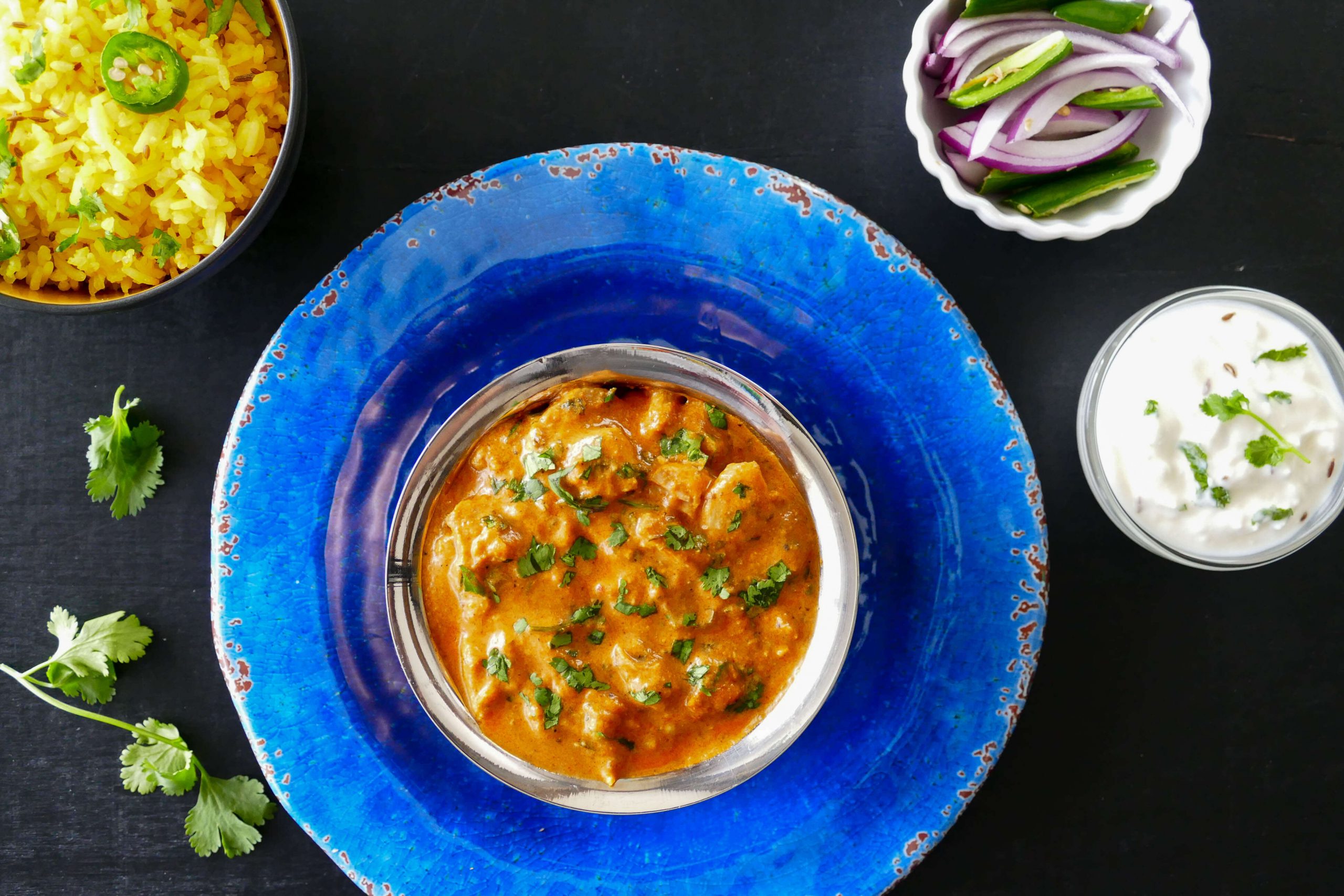 Instant Pot Chicken Tikka Masala in bowl on bright blue plate, with yellow rice, red onions and green chilis and yogurt (raita) in bowls in background - Paint the Kitchen Red