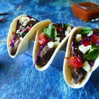 Instant Pot Carne Asada - three tacos with purple cabbage, tomatoes, cheese, cilantro on a blue background with salsa, red chilis and cut lime