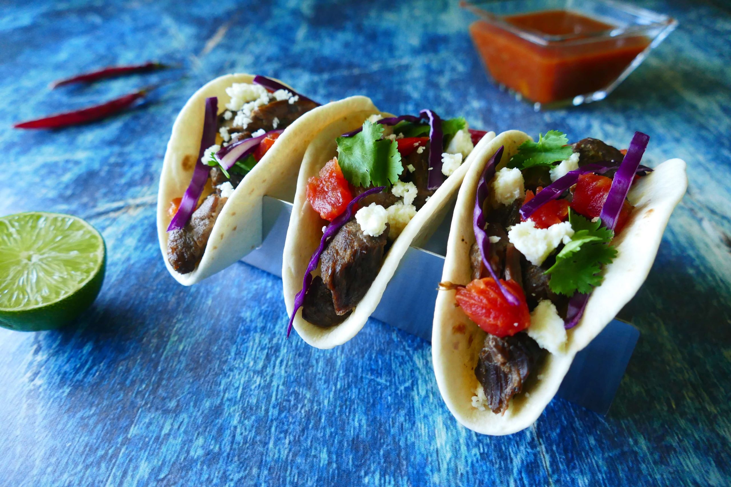 Instant Pot Carne Asada - three tacos with purple cabbage, tomatoes, cheese, cilantro on a blue background with salsa, red chilis and cut lime