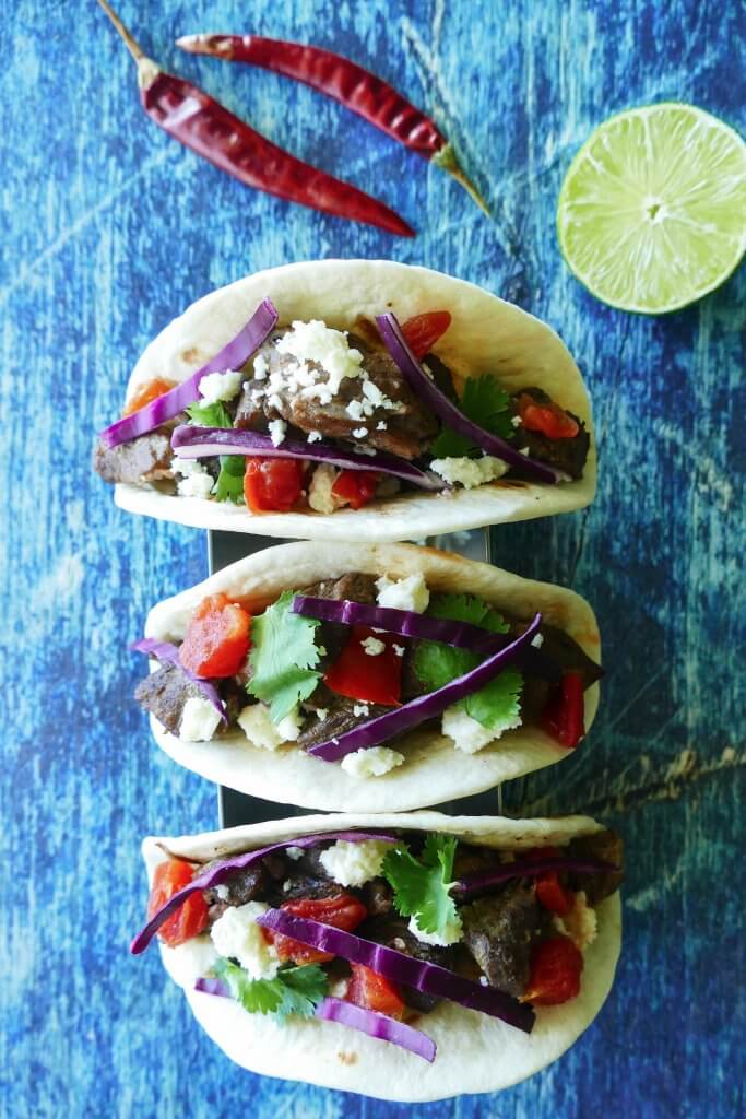 Instant Pot Carne Asada - three tacos with purple cabbage, tomatoes, cheese on a blue background with red chilis and cut lime