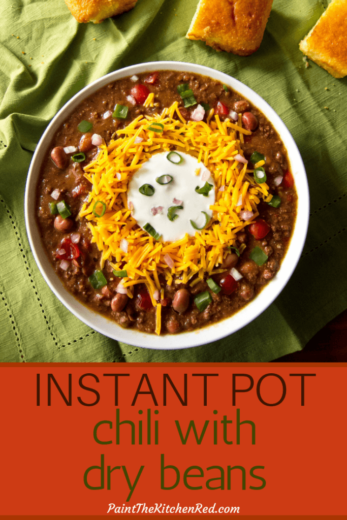 Instant Pot Chili Pinterest pin with image of chili with shredded cheddar surrounding sour cream, topped with green onions. On a green cloth with pieces of corn bread. From Paint the Kitchen Red