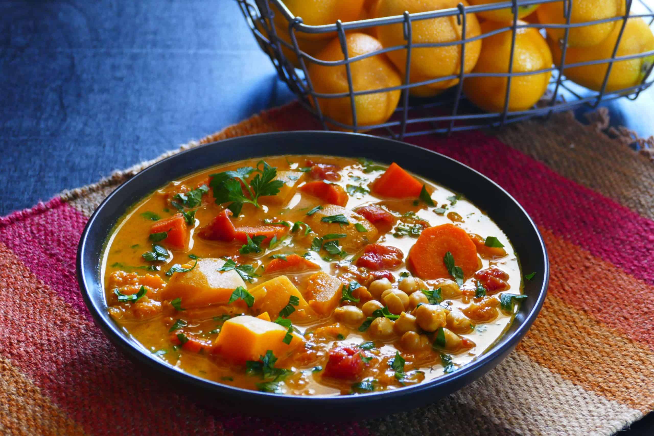 Vegetarian Instant Pot Moroccan Stew with carrots, squash, chickpeas, cilantro, tomatoes in black bowl on colorful mat with lemons in background