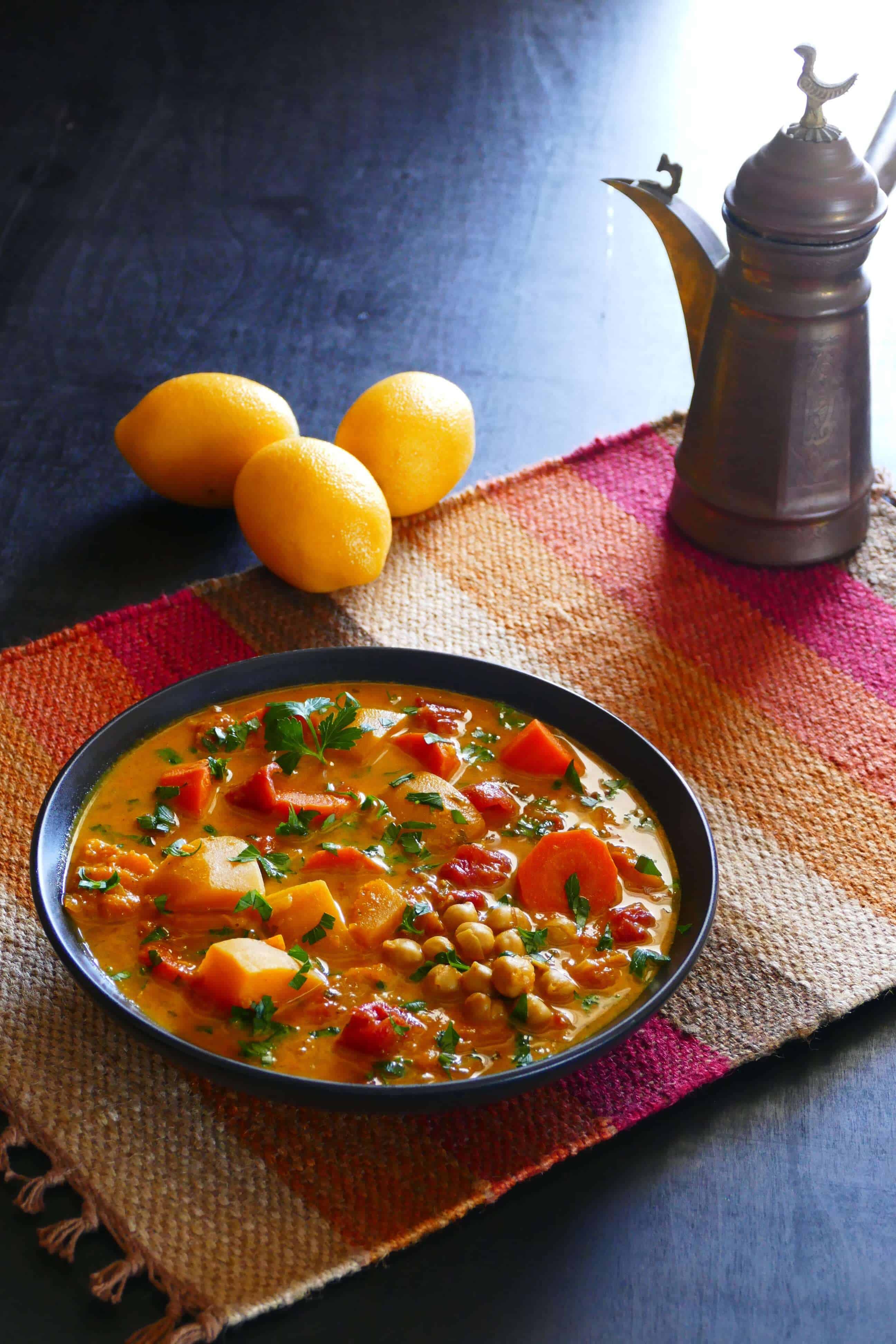 Vegetarian Instant Pot Moroccan Stew with carrots, squash, chickpeas, cilantro, tomatoes in black bowl on colorful mat with lemons in background