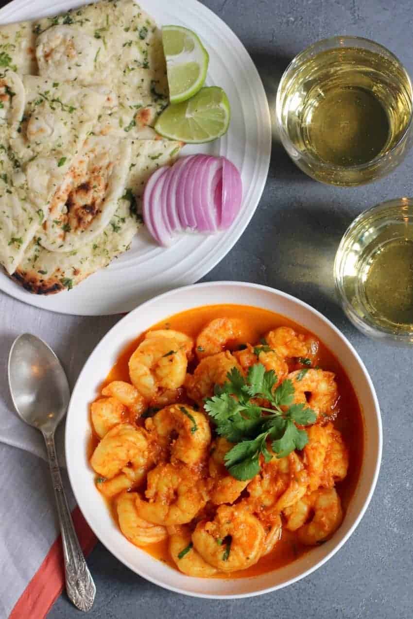 Bowl of shrimp curry garnished with cilantro with a plate of naan in the background