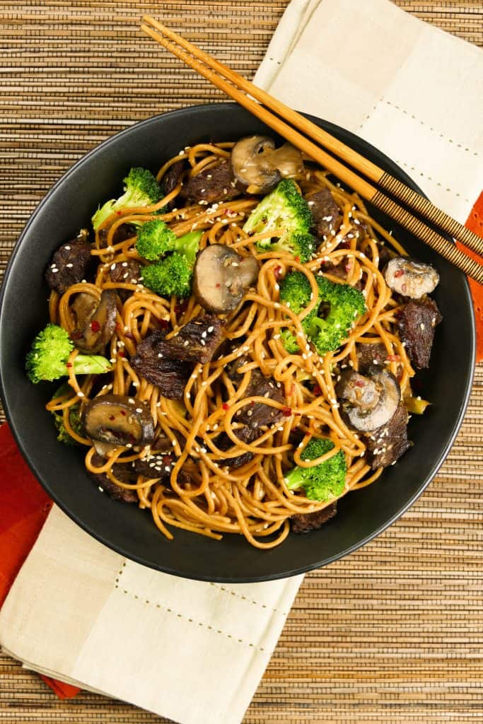 Instant Pot Lo Mein with Beef and Broccoli in black bowl on straw mat with chopsticks laid across bowl - Paint the Kitchen Red