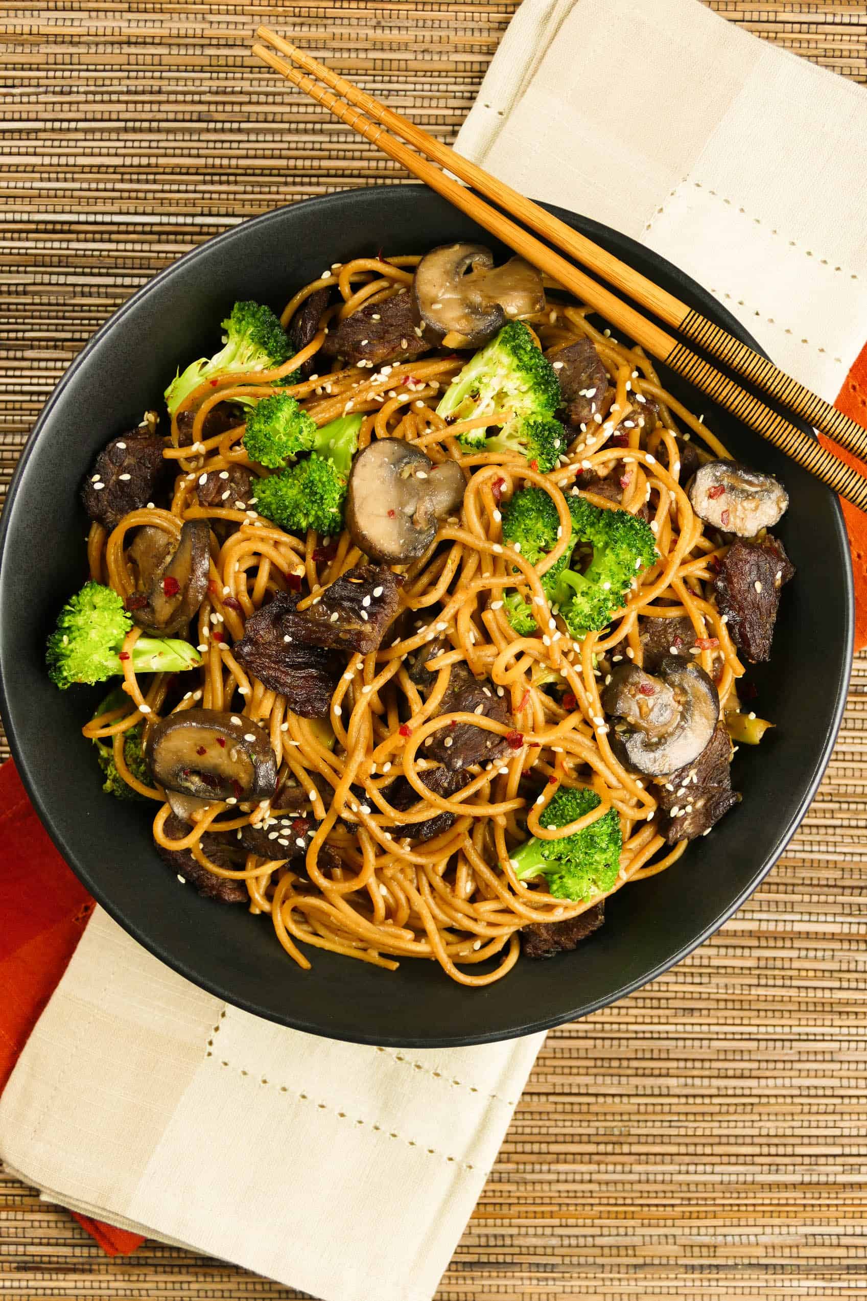 Instant Pot Lo Mein with Beef and Broccoli in black bowl on straw mat with chopsticks laid across bowl - Paint the Kitchen Red