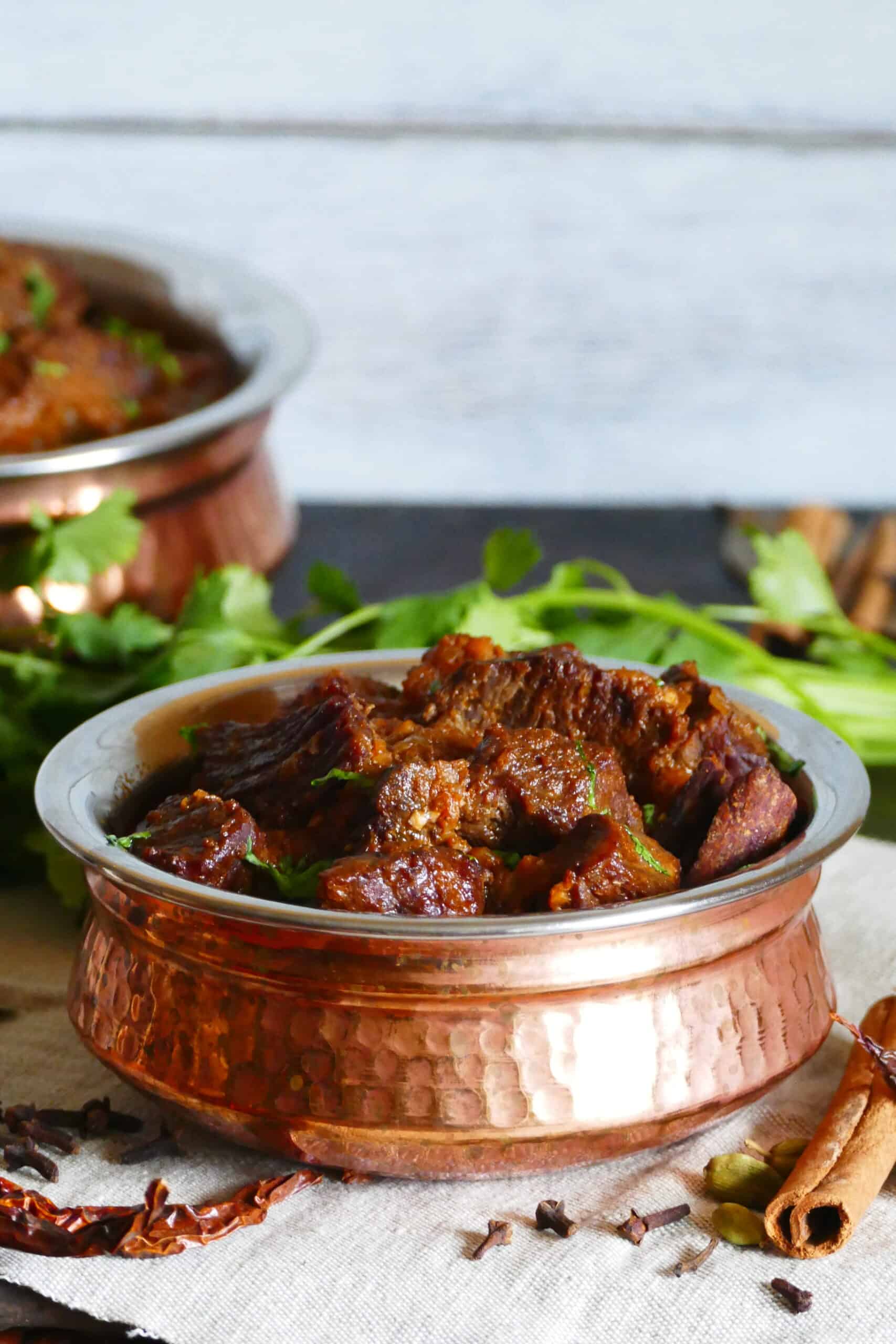 Instant Pot Rogan Josh in a copper dish with whole spices and cilantro in background