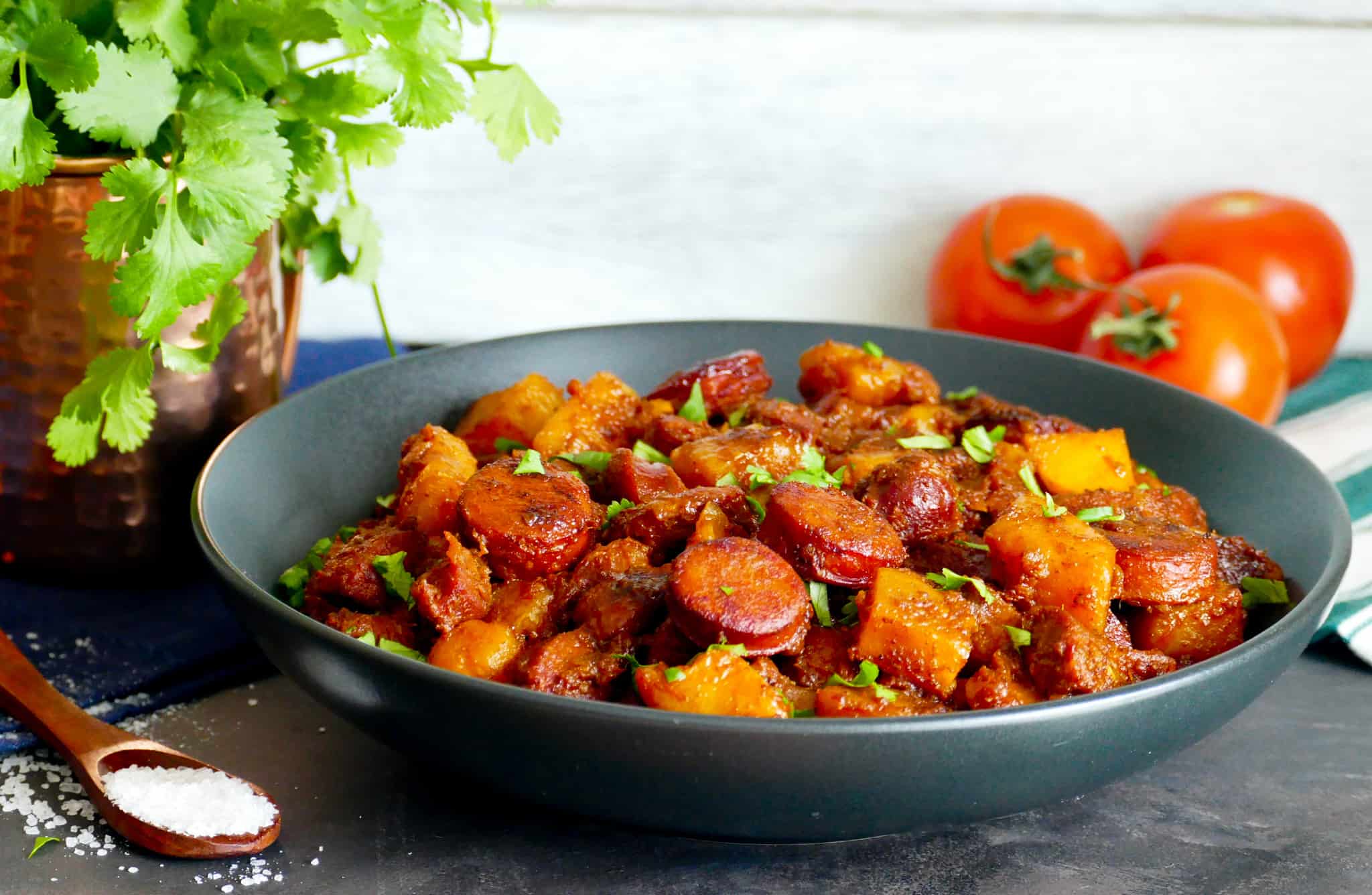 Black round serving dish with chorizo and potato sprinkled with cilantro and copper mug with fresh cilantro and fresh tomatoes in background