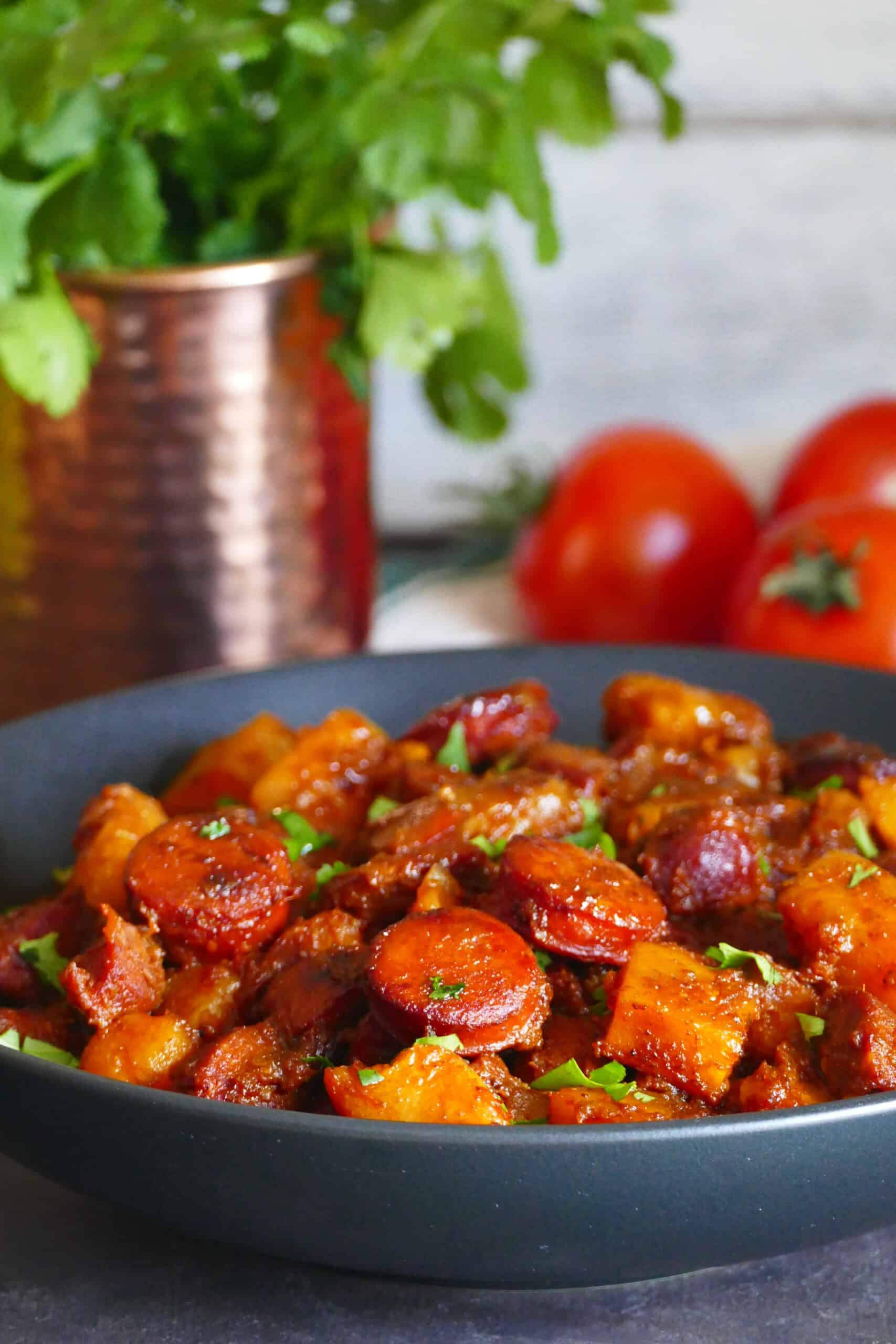 Black round serving dish with chorizo and potato sprinkled with cilantro and copper mug with fresh cilantro and fresh tomatoes in background