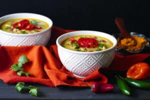 Two white bowls of yellow colored chicken curry soup garnished with red jalapenos and green onions on an orange napkin