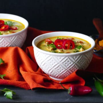 Two white bowls of yellow colored chicken curry soup garnished with red jalapenos and green onions on an orange napkin