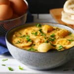 Blue speckled bowl of egg curry garnished with cilantro, with naan and brown eggs in the background