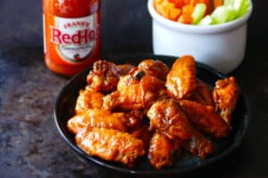 Blue plate with hot wings glistening with sauce and carrots, celery in a white bowl and Frank's redhot sauce in background