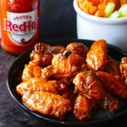 Blue plate with hot wings glistening with sauce and carrots, celery in a white bowl and Frank's redhot sauce in background