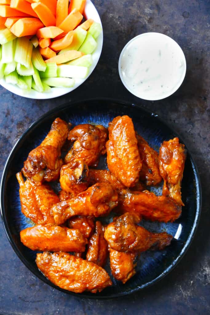 Blue plate with hot wings glistening with sauce and carrots, celery in a white bowl and ranch sauce in a bowl