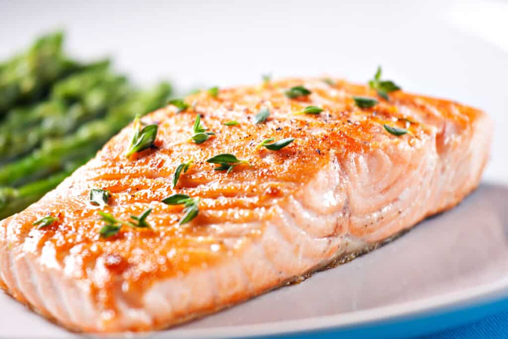 filet of salmon on a white plate garnished with herbs and asparagus in the background