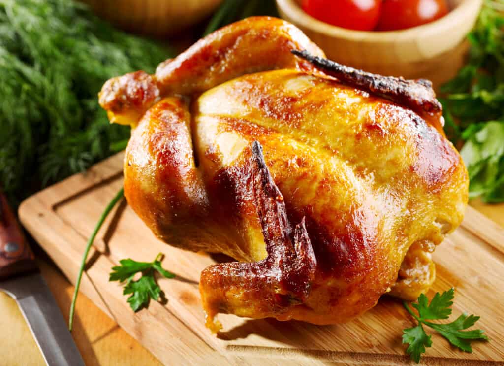 whole chicken on a cutting board surrounded by greens and tomatoes in a bowl