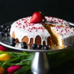Pound cake on cake stand frosted with white frosting and topped with red balls and a ripe strawberry and blue berries on the side