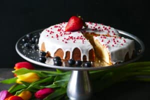Pound cake on cake stand frosted with white frosting and topped with red balls and a ripe strawberry and blue berries on the side