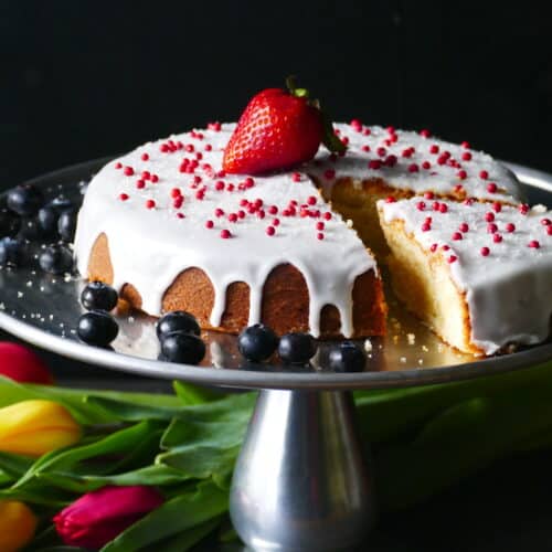 Pound cake on cake stand frosted with white frosting and topped with red balls and a ripe strawberry and blue berries on the side