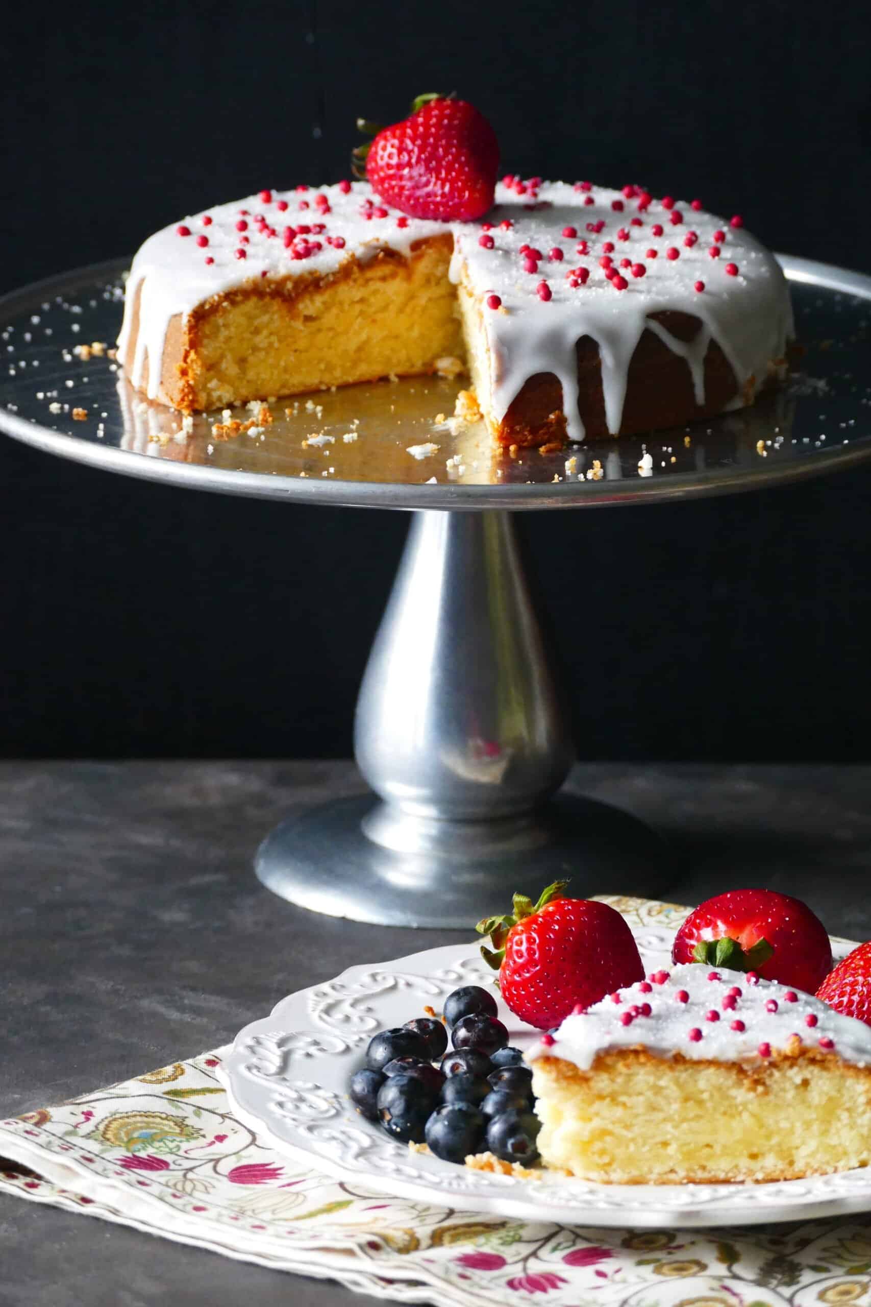 Air fryer cake on cake stand frosted with white frosting and topped with red balls and a ripe strawberry