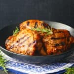 Grilled peri peri chicken pieces stacked in black plate on blue and white napkin with rosemary garnish