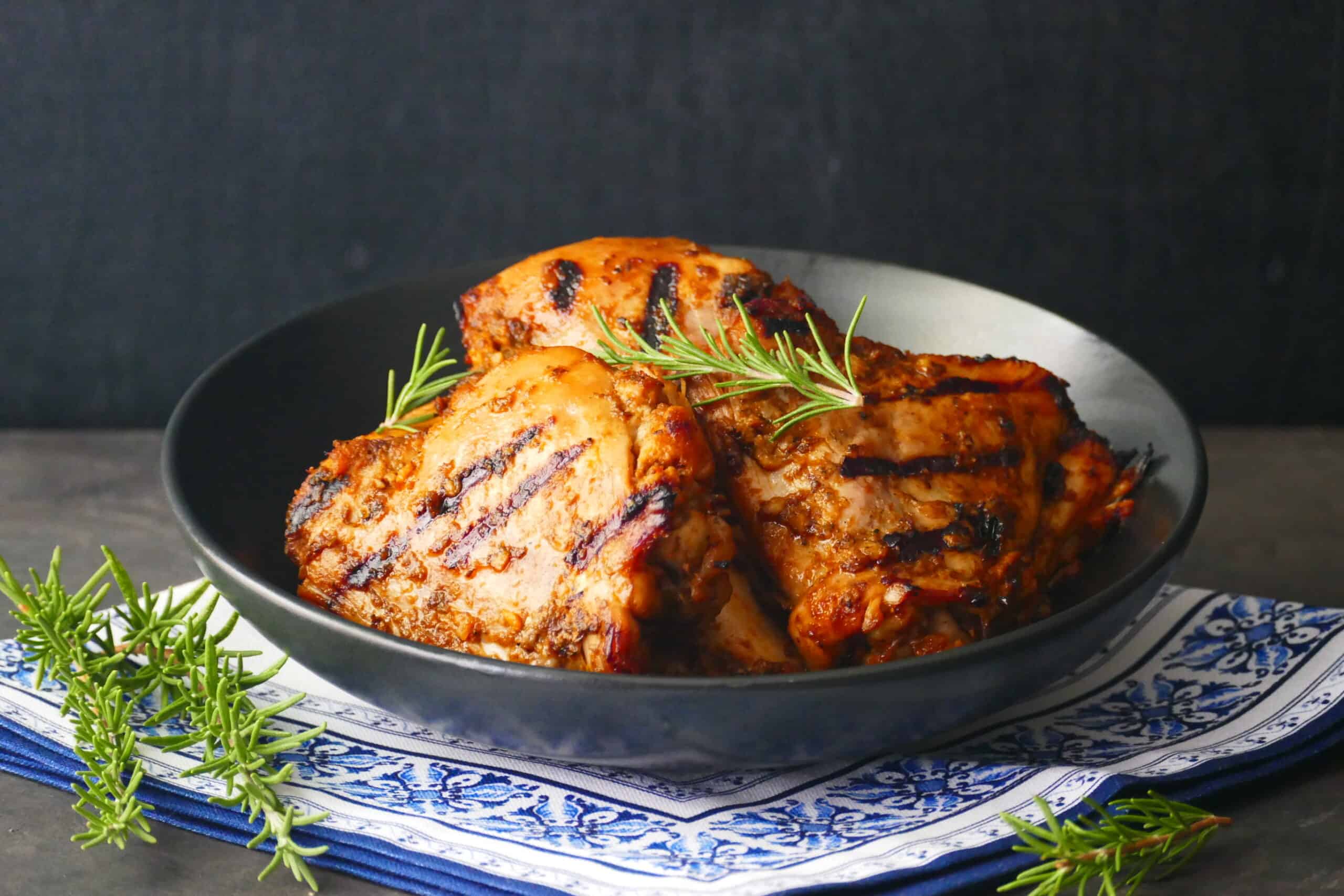 Grilled peri peri chicken pieces stacked in black plate on blue and white napkin with rosemary garnish