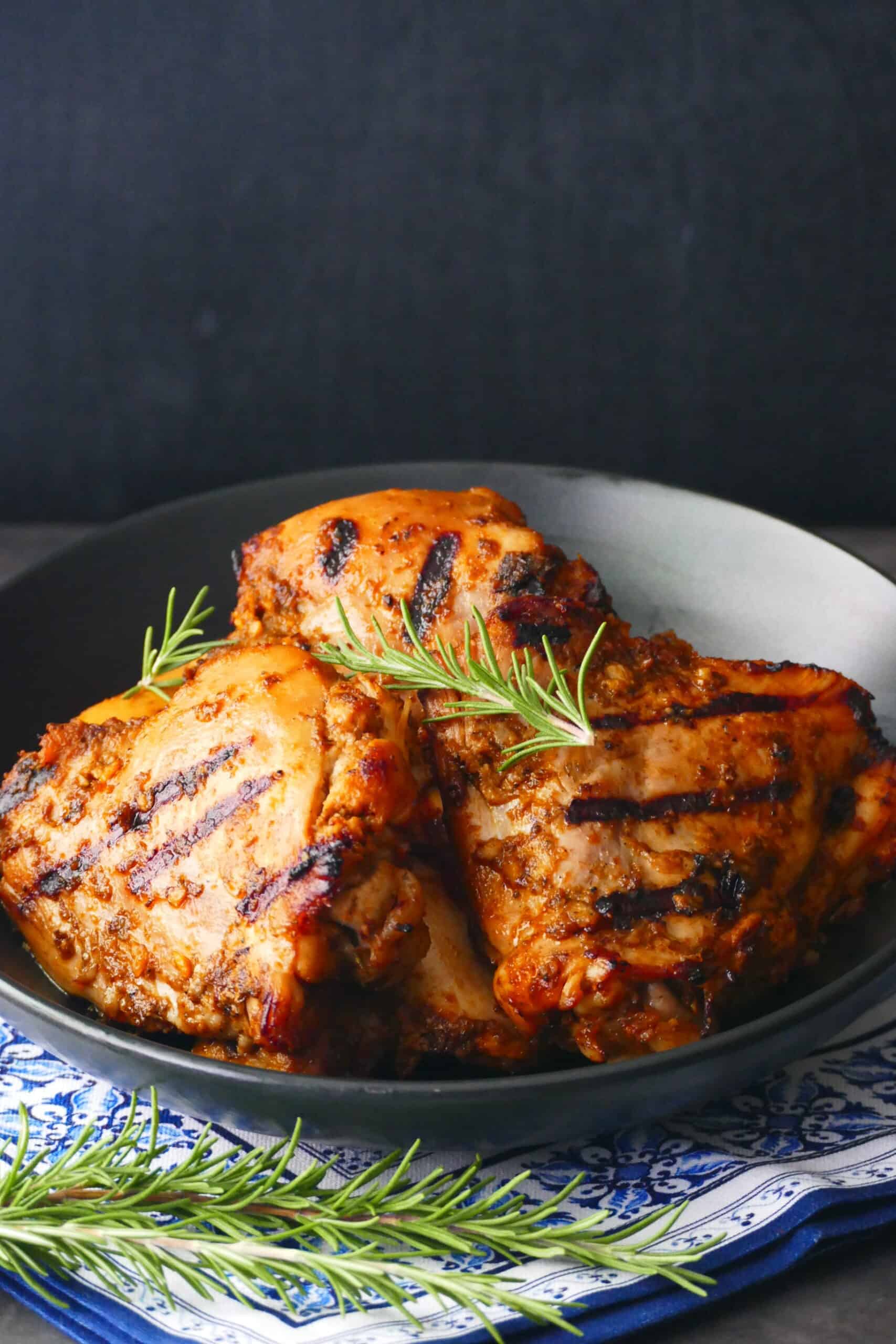 Grilled peri peri chicken pieces stacked in black plate on blue and white napkin with rosemary garnish