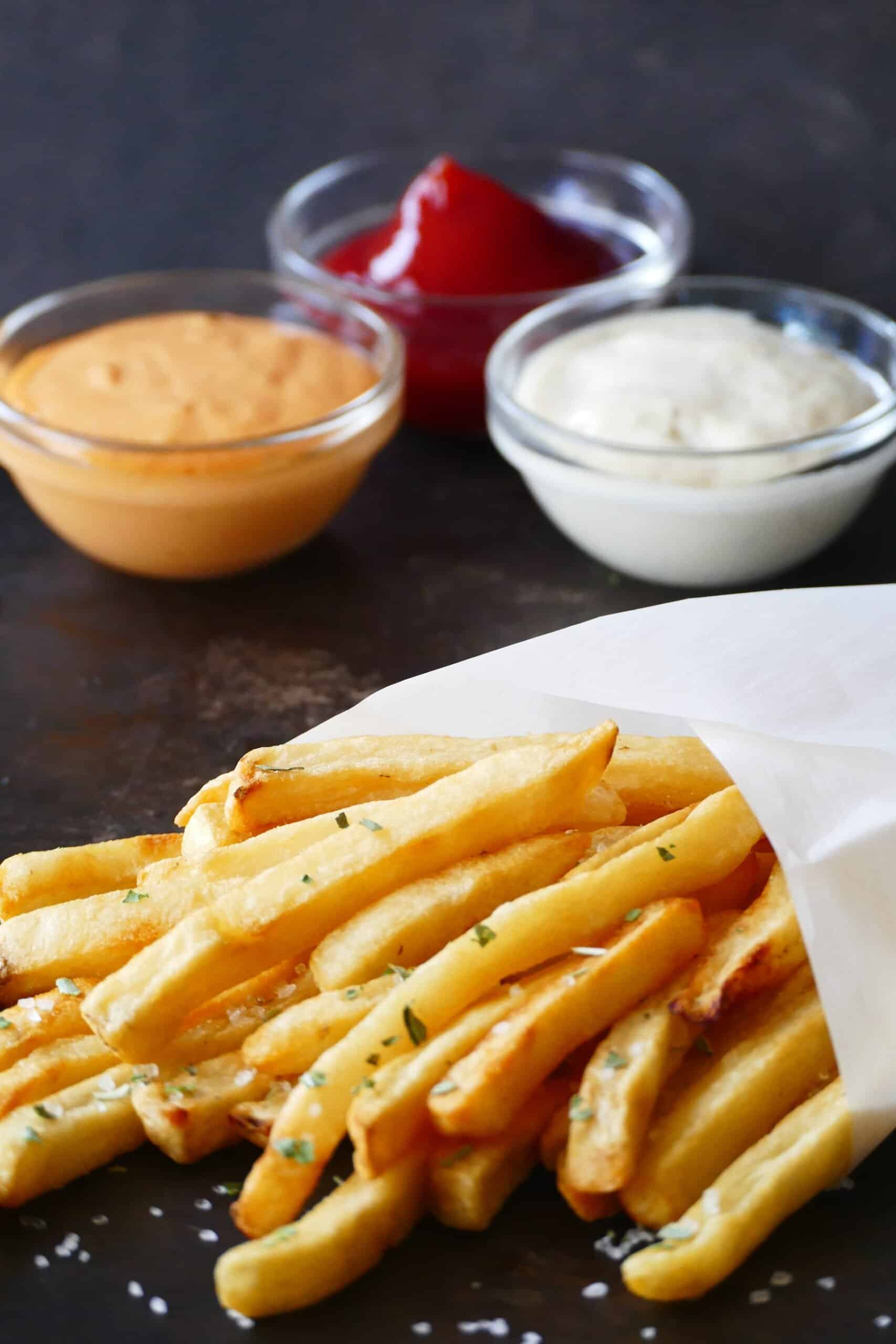 Instant Pot air fryer frozen french fries with sea salt and parsley wrapped in white parchment and three glass bowls with condiments