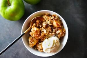 White bowl with apple crisp and scoop of ice cream with a gold spoon nestled in the bowl and green apple in the background