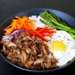 Black bowl with pulled pork, carrots, red onions, bell pepper, asparagus, fried egg and rice on black background