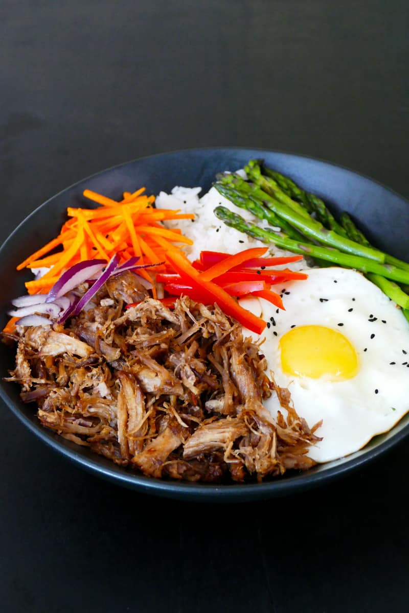 Black bowl with pulled pork, carrots, red onions, bell pepper, asparagus, fried egg and rice on black background